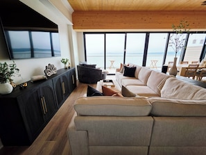 A modern living room with a large sofa, a flat-screen TV mounted on the wall, and a view of the sea through large windows.