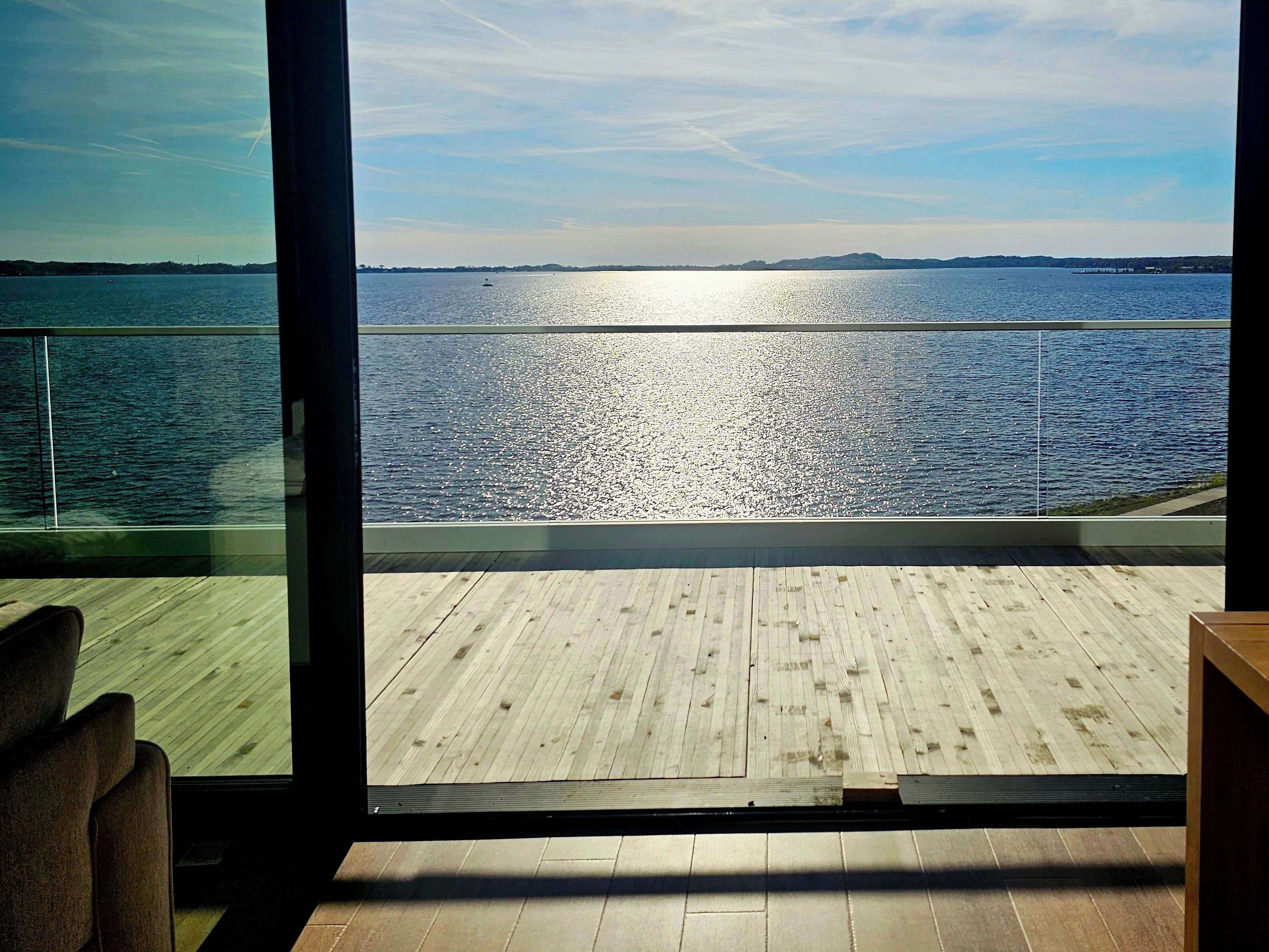 Una terraza de madera con barandilla de cristal que da a una masa de agua tranquila.