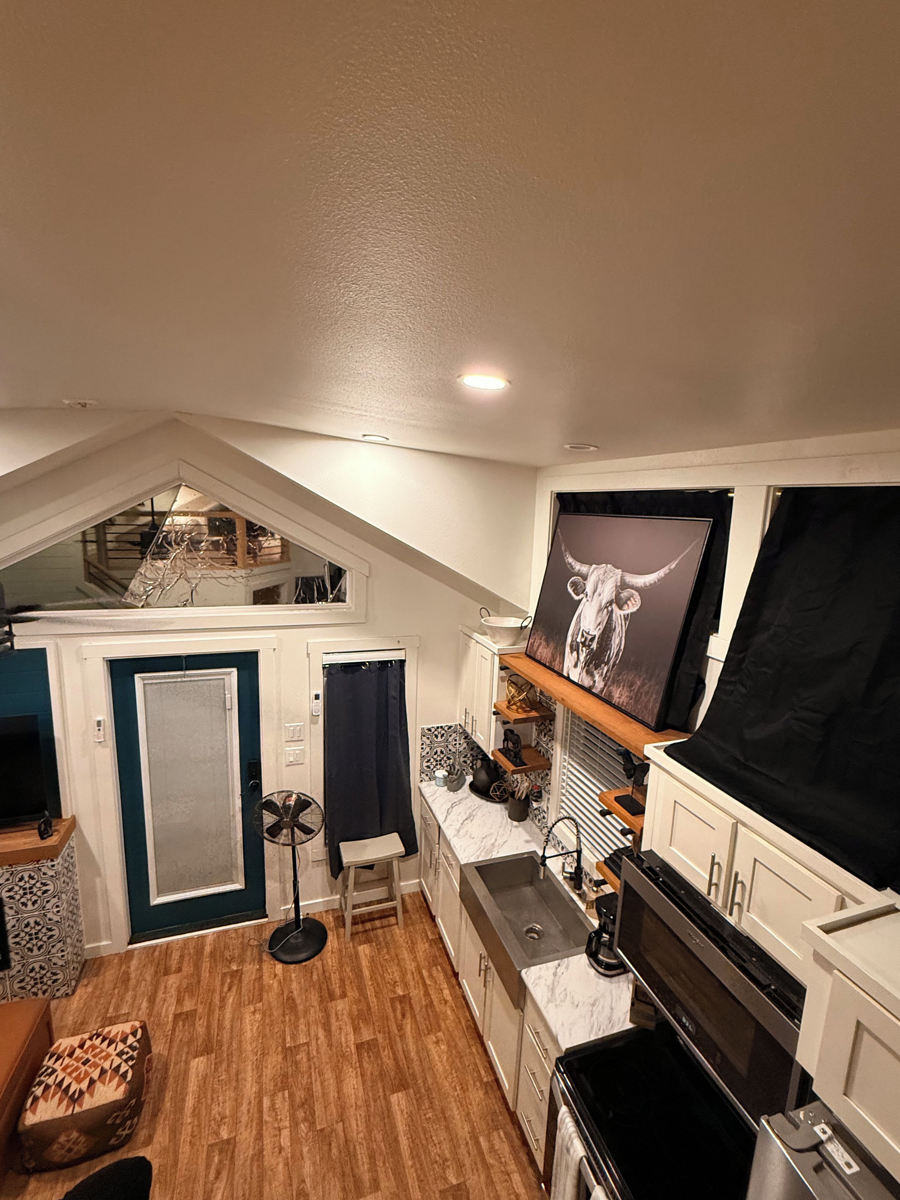 A compact kitchen with a microwave, stove, and sink. There is a small stool and a fan. The room has a sloped ceiling and a window with a curtain.