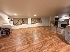 A spacious attic room with wooden flooring, two windows with curtains, a white pillow, and a fan.