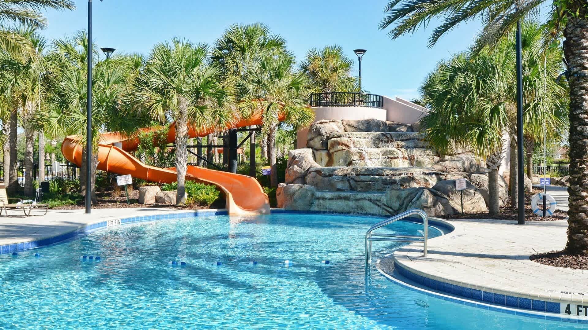 A swimming pool with a water slide, palm trees, and a rocky waterfall feature.