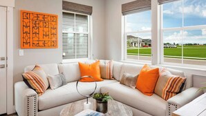Un salon moderne avec un canapé d’angle beige, une table basse ronde et une grande fenêtre donnant sur un quartier résidentiel.