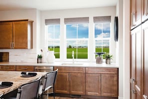 A kitchen with wooden cabinets, a large window with a view of a green field, and a dining area with a table and chairs.