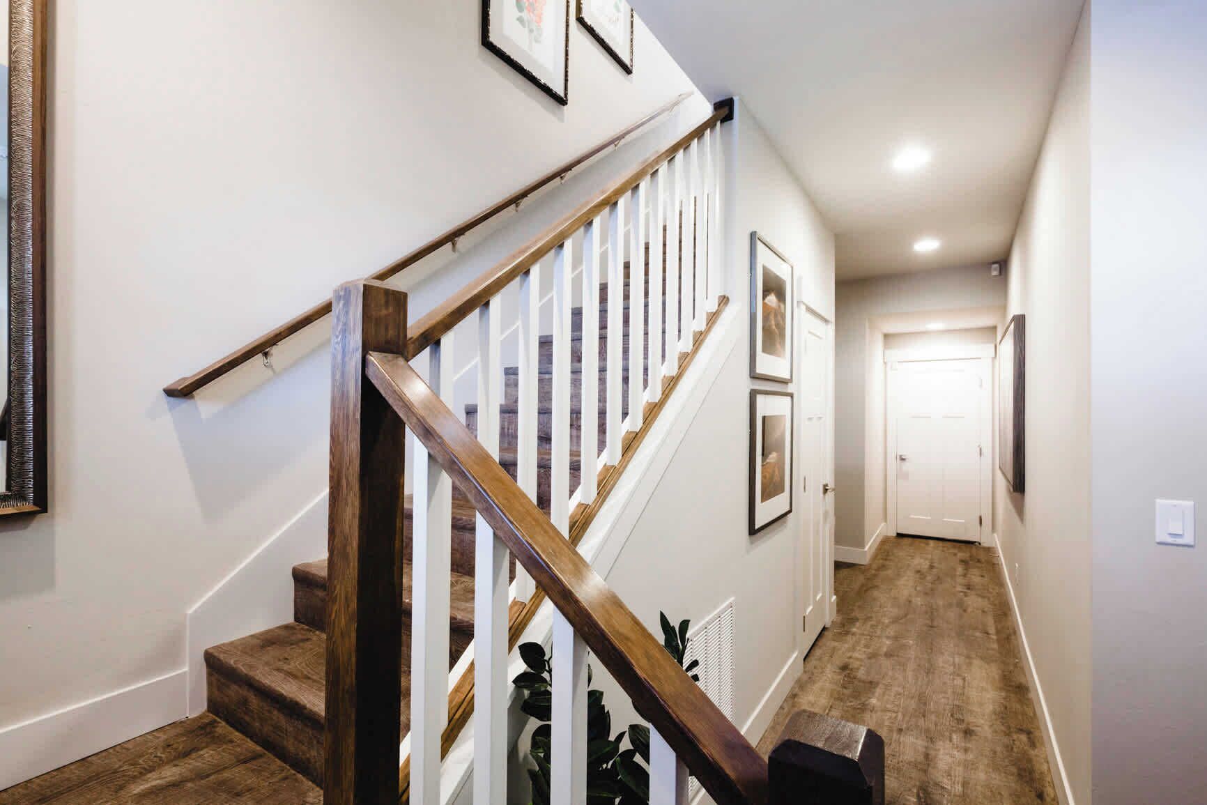 A staircase with wooden handrails and white balusters, leading to a door at the end of a hallway with framed pictures on the walls.