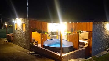 Un área de hidromasaje con revestimiento de piedra, rodeada por una terraza de madera e iluminada por luces exteriores.