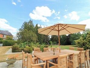 Ein Garten mit einem Holztisch, Stühlen und einem großen Sonnenschirm, im Hintergrund ein Haus und ein Hecke.