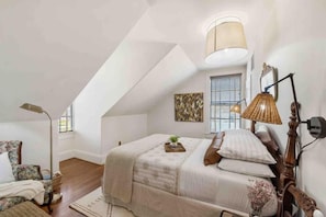 A bedroom with a sloped ceiling, a bed with a headboard, a floor lamp, a chair, and a window with blinds. - Rustic Luxury from Restoring Galveston (Galveston)