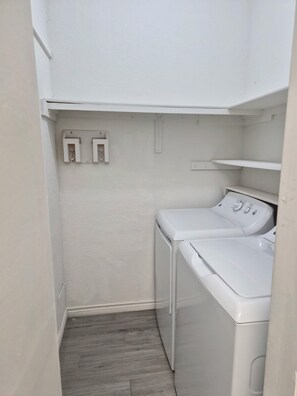 A laundry room with two white washing machines and a dryer, a wooden floor, and a white wall with electrical outlets. - I114 – Private King Retreat | DFW 10 Min | Yard+W/D (Irving)