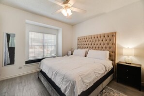 Een slaapkamer met een bed, een plafondventilator, een raam met zonwering, een nachtkastje met een lamp en een vloerkleed.