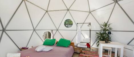 A geodesic dome room with a bed, pillows, a hanging chair, a small table, a plant, and a heater.