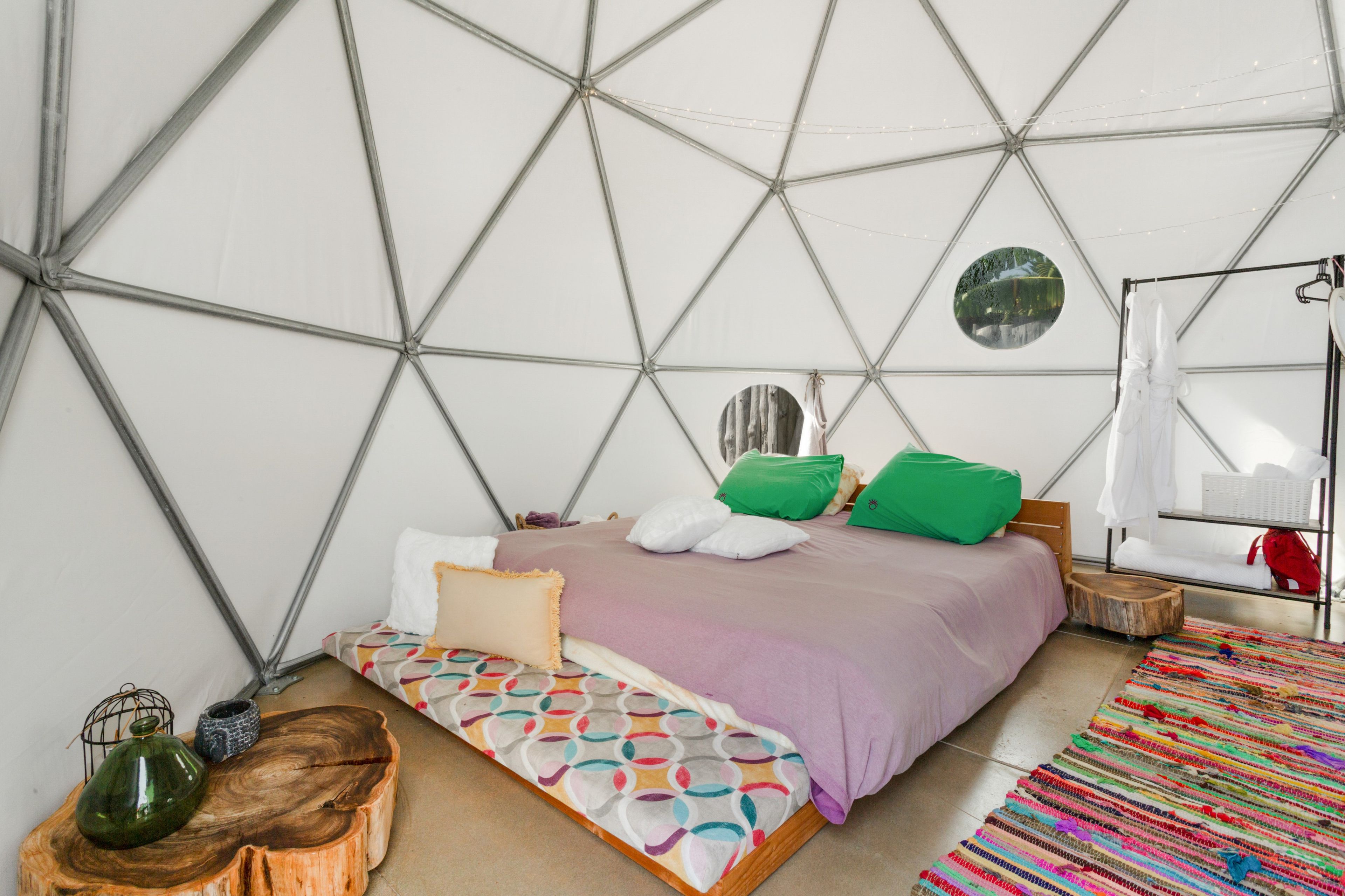 A room with a geometric ceiling structure, a bed with a colorful patterned bedspread, a small wooden bench, and a colorful woven rug.