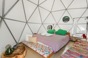 Ein Zimmer mit einer geometrischen Deckenstruktur, einem Bett mit einer farbenfrohen Musterdecke, einer kleinen Holzbank und einem farbenfrohen Teppich.