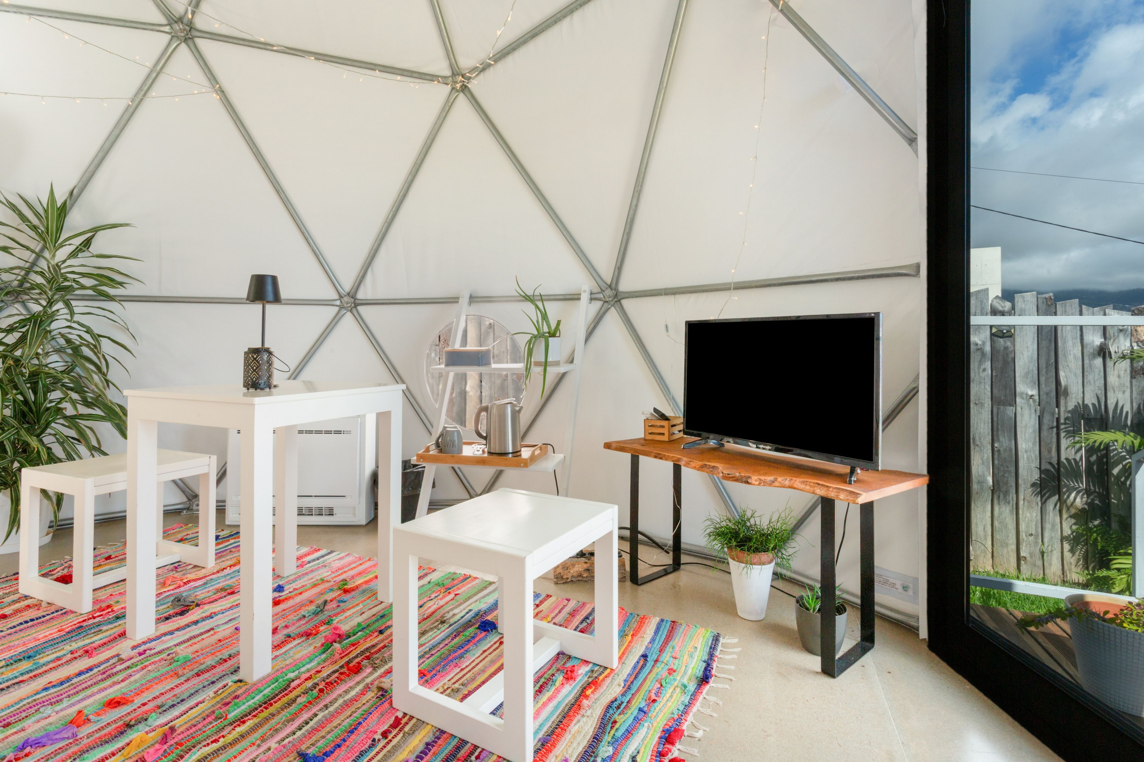 Een moderne woonruimte met een geometrisch plafond, een flatscreen televisie, een salontafel, een klein tafeltje met een lamp, en een kleurrijk vloerkleed.