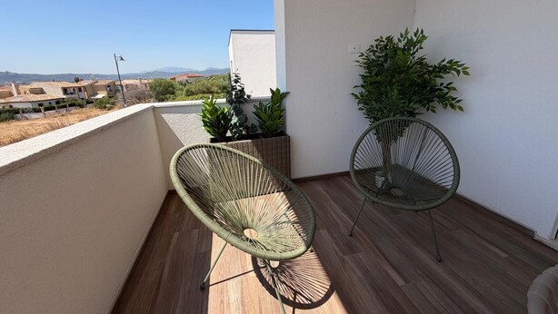 A balcony with two wicker chairs, a potted plant, and a view of distant buildings. - Le Calendule Luxury Apartment (Olbia)