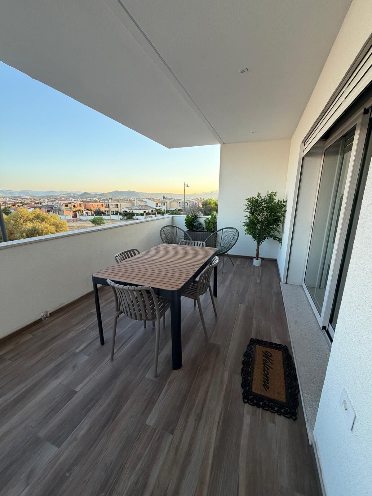 A balcony with a wooden table and chairs, a potted plant, and a view of a cityscape.