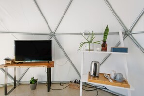 Ett rum med en tv på ett träbord, en krukväxt, en hylla med en vattenkokare, en mugg och en sked.
