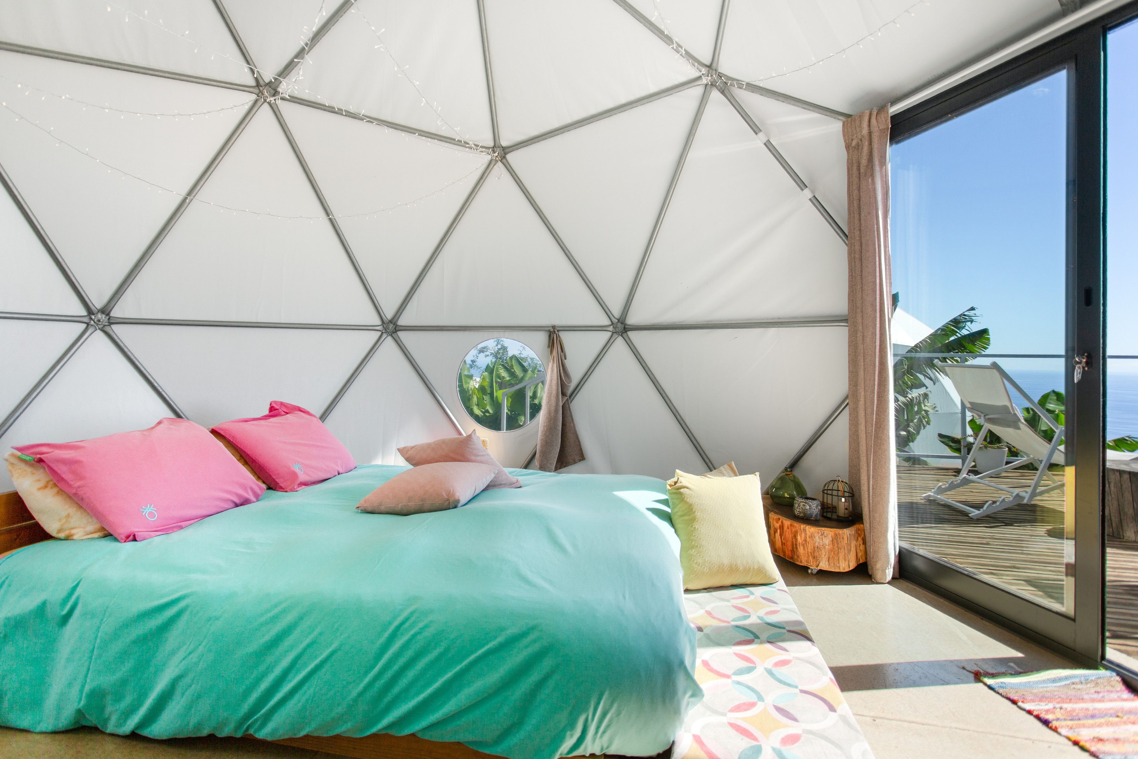 Een moderne slaapkamer met een geometrisch plafondontwerp, een bed met veel kussens, een klein rond spiegelbeeld en uitzicht op de omgeving door een schuifdeur van glas.
