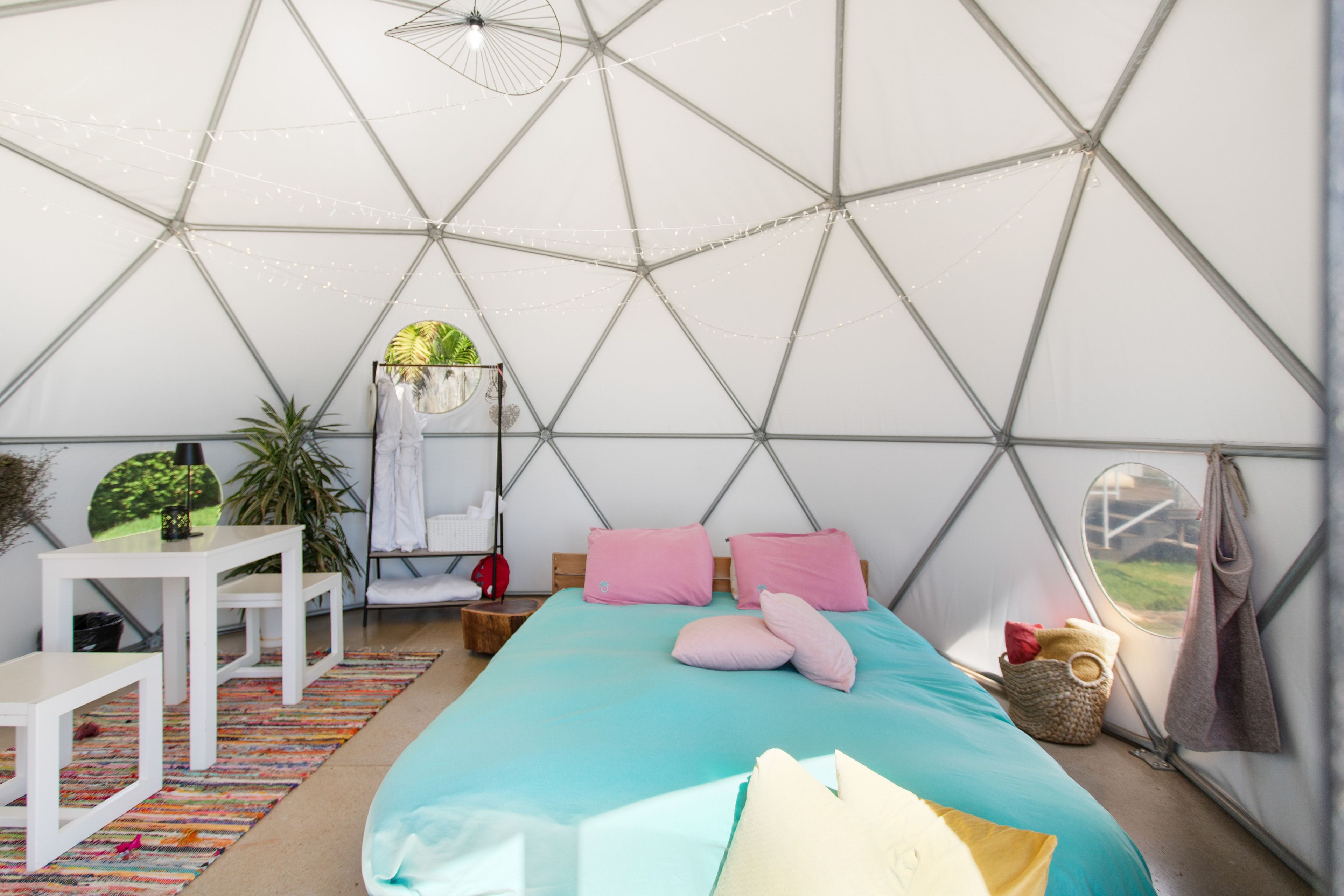 Eine Zelthütte mit einem Bett, Kissen, einem Hängesessel, einem Tisch und einer Pflanze.