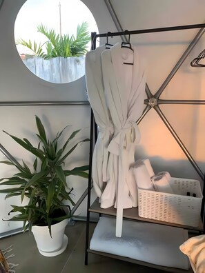 A room with a white wall, a round window, a white potted plant, a white robe hanging on a metal rack, and a white basket on a shelf.