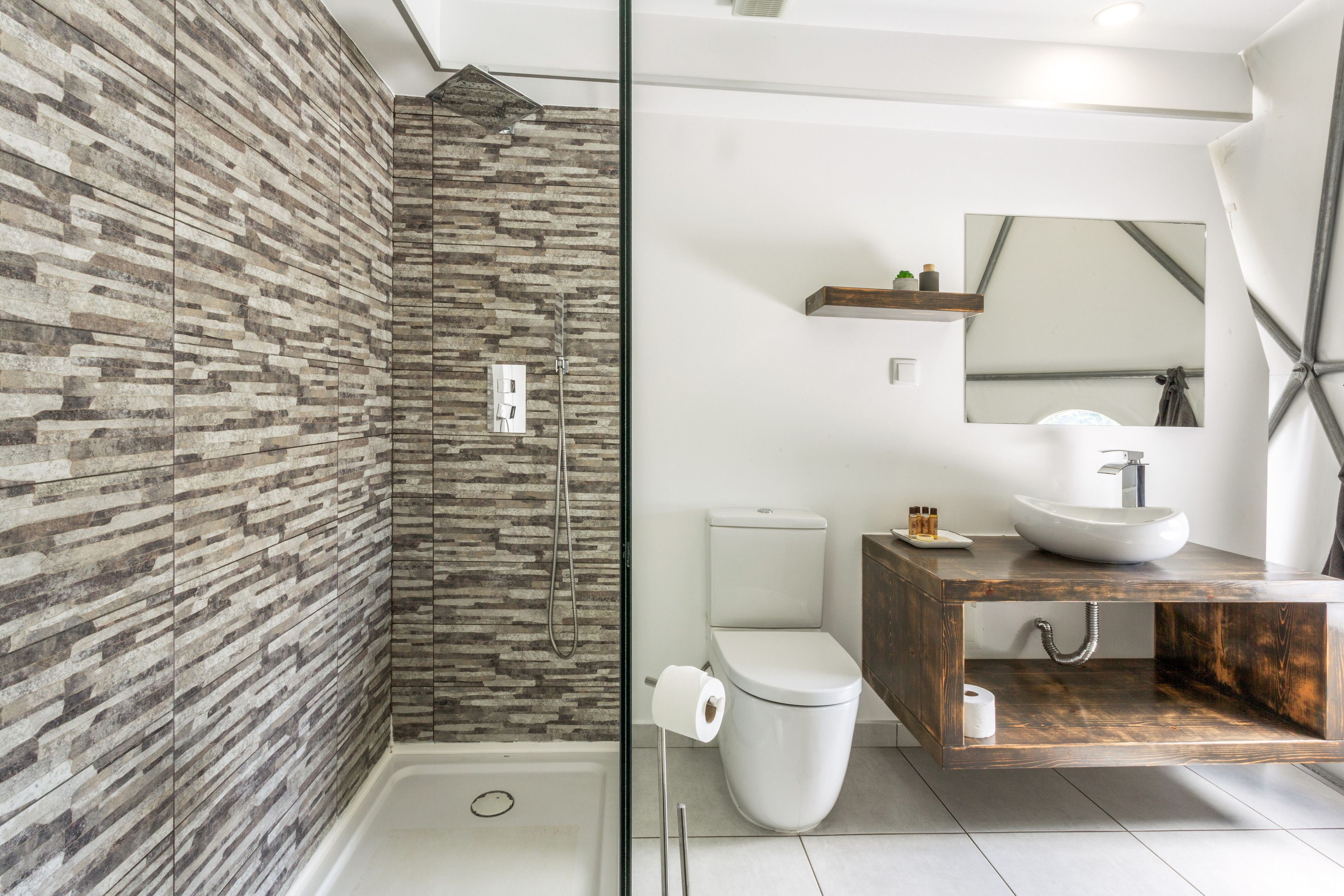 Un baño moderno con pared de piedra, ducha envuelta en vidrio, inodoro blanco y tocador de madera con lavamanos.