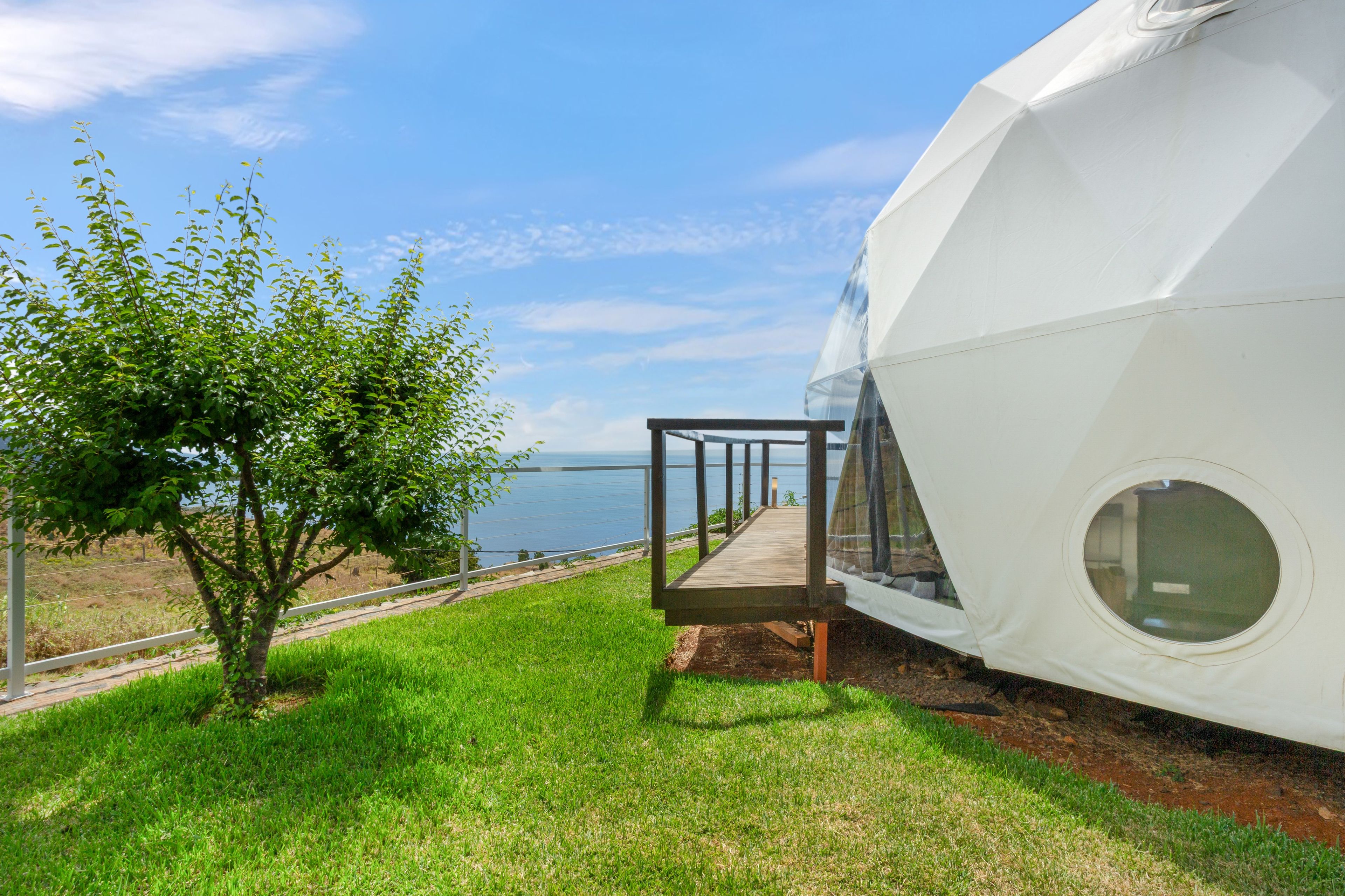 A modern, white, dome-shaped building with a wooden deck leading to a glass door, overlooking a body of water.