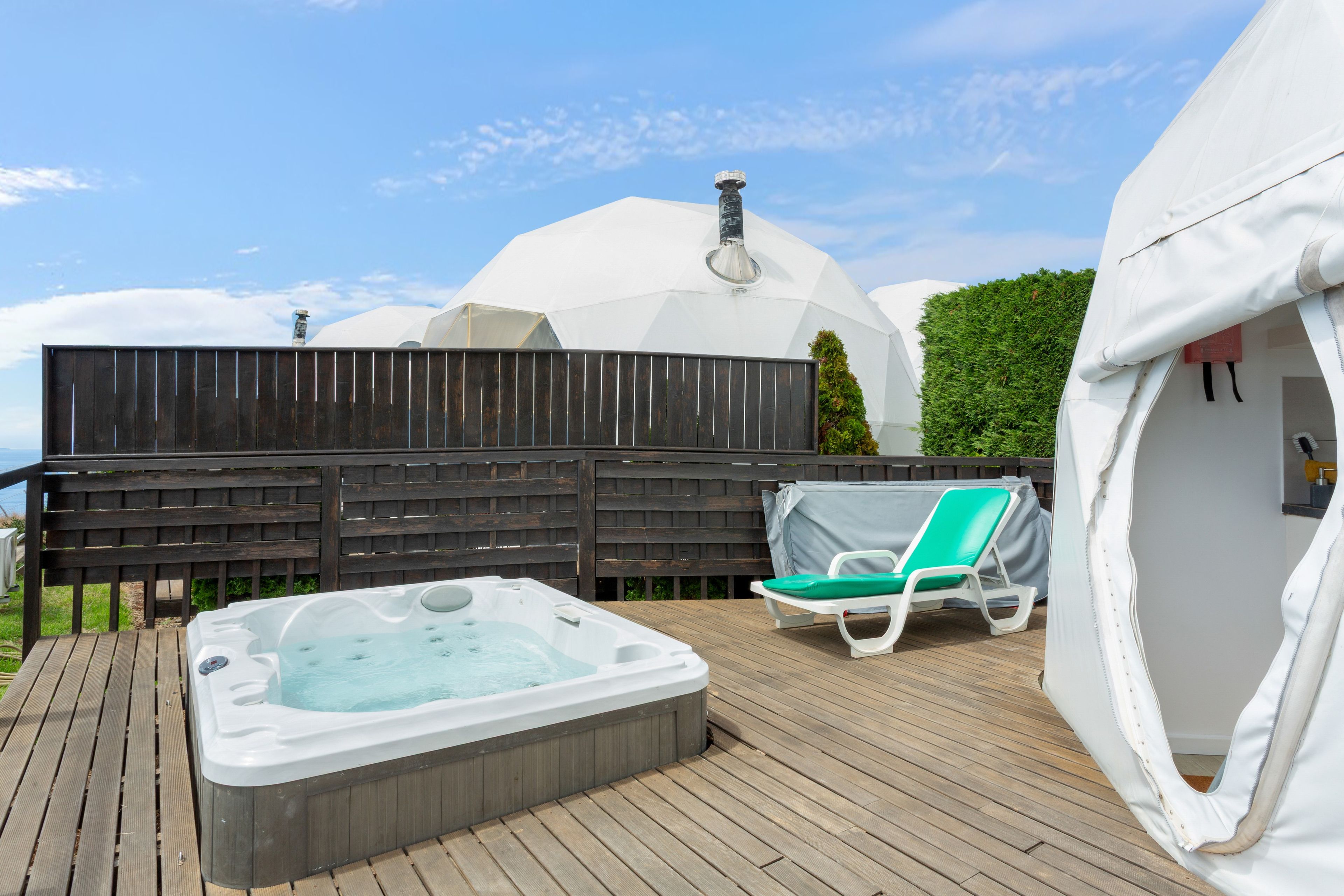 A hot tub on a wooden deck with a covered lounge chair and a dome-shaped structure in the background.
