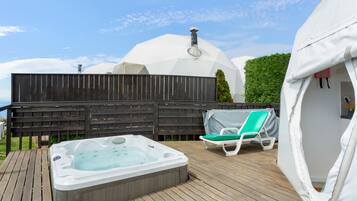 A hot tub on a wooden deck with a covered lounge chair and a dome-shaped structure in the background.
