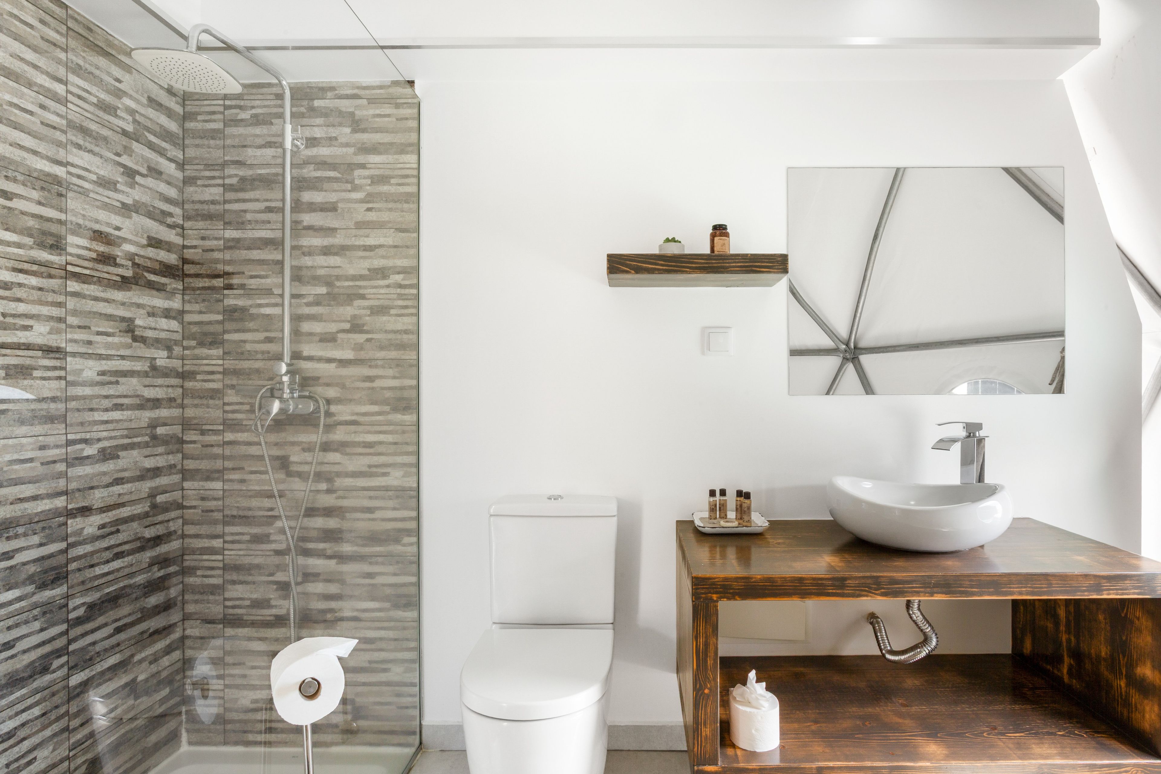 A modern bathroom with a glass shower enclosure, a white toilet, a wooden vanity with a vessel sink, and a small shelf with toiletries.
