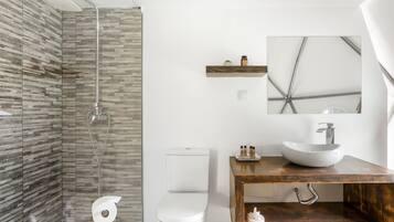 A modern bathroom with a glass shower enclosure, a white toilet, a wooden vanity with a vessel sink, and a small shelf with toiletries.