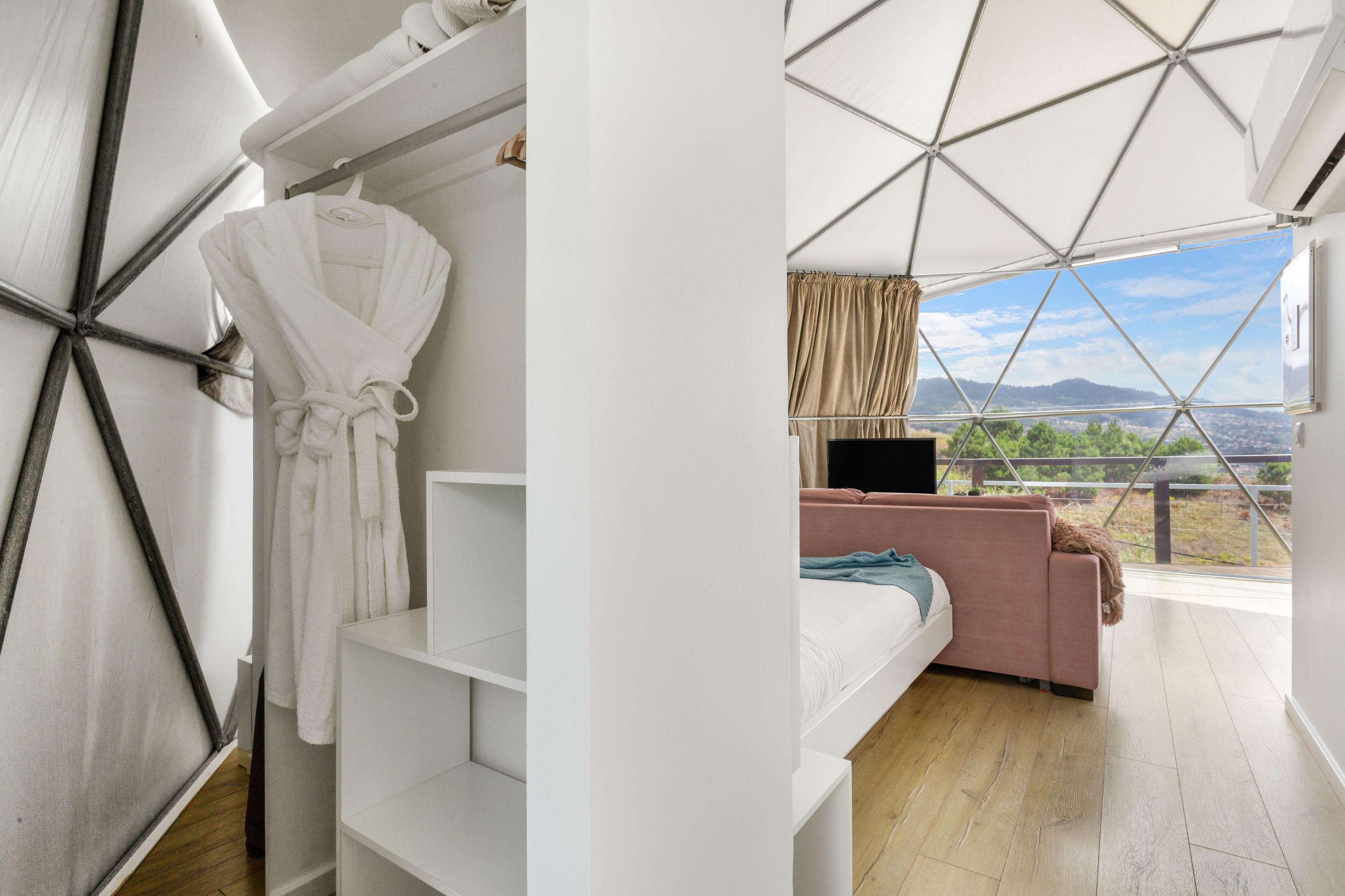 Ein modernes Zimmer mit einem Etagenbett, einem Kleiderhaken mit einem Bademantel, einer Couch und einem großen Fenster mit Blick ins Freie.