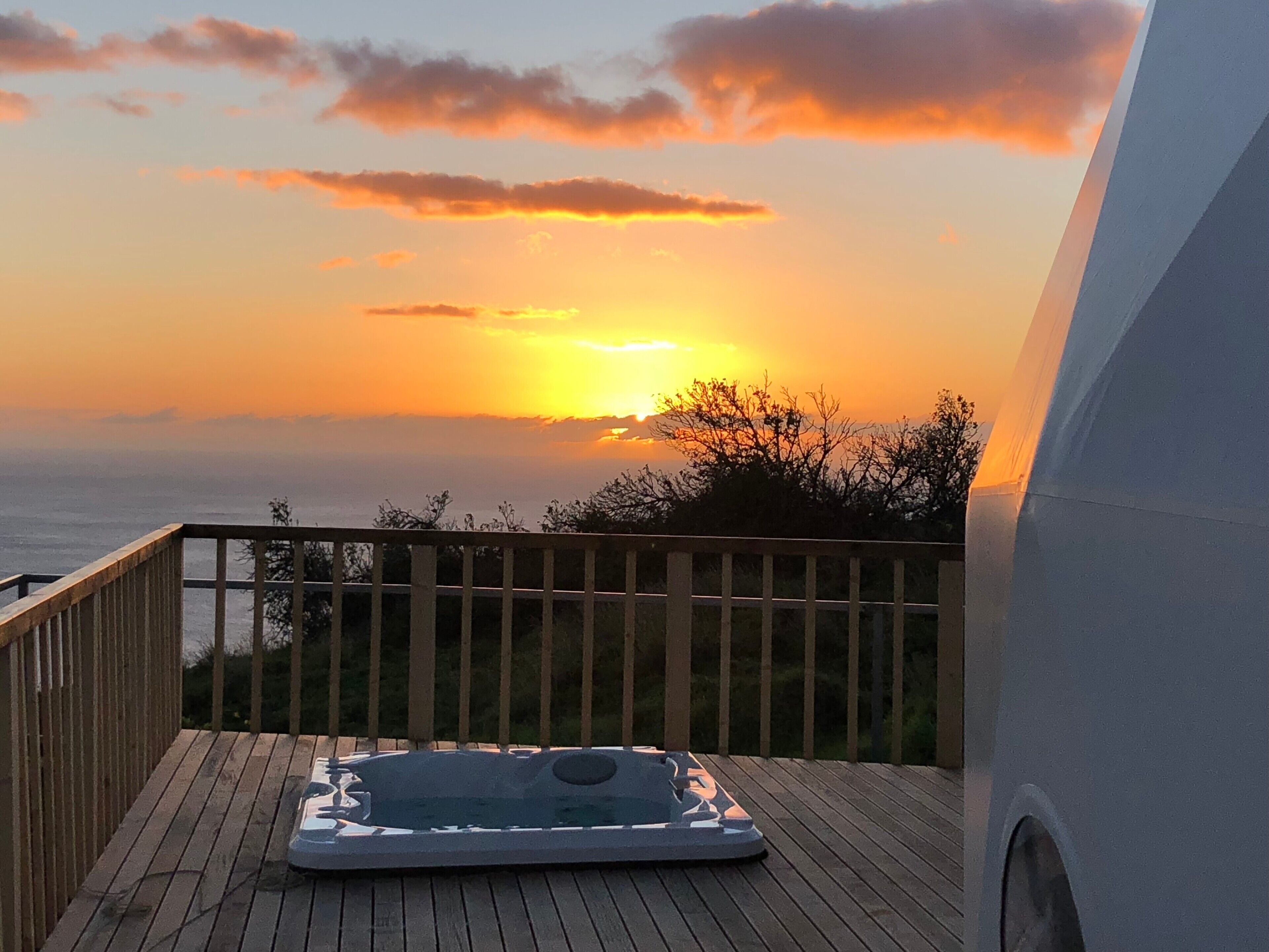 Een houten terras met een jacuzzi en uitzicht op de ondergaande zon boven de oceaan.