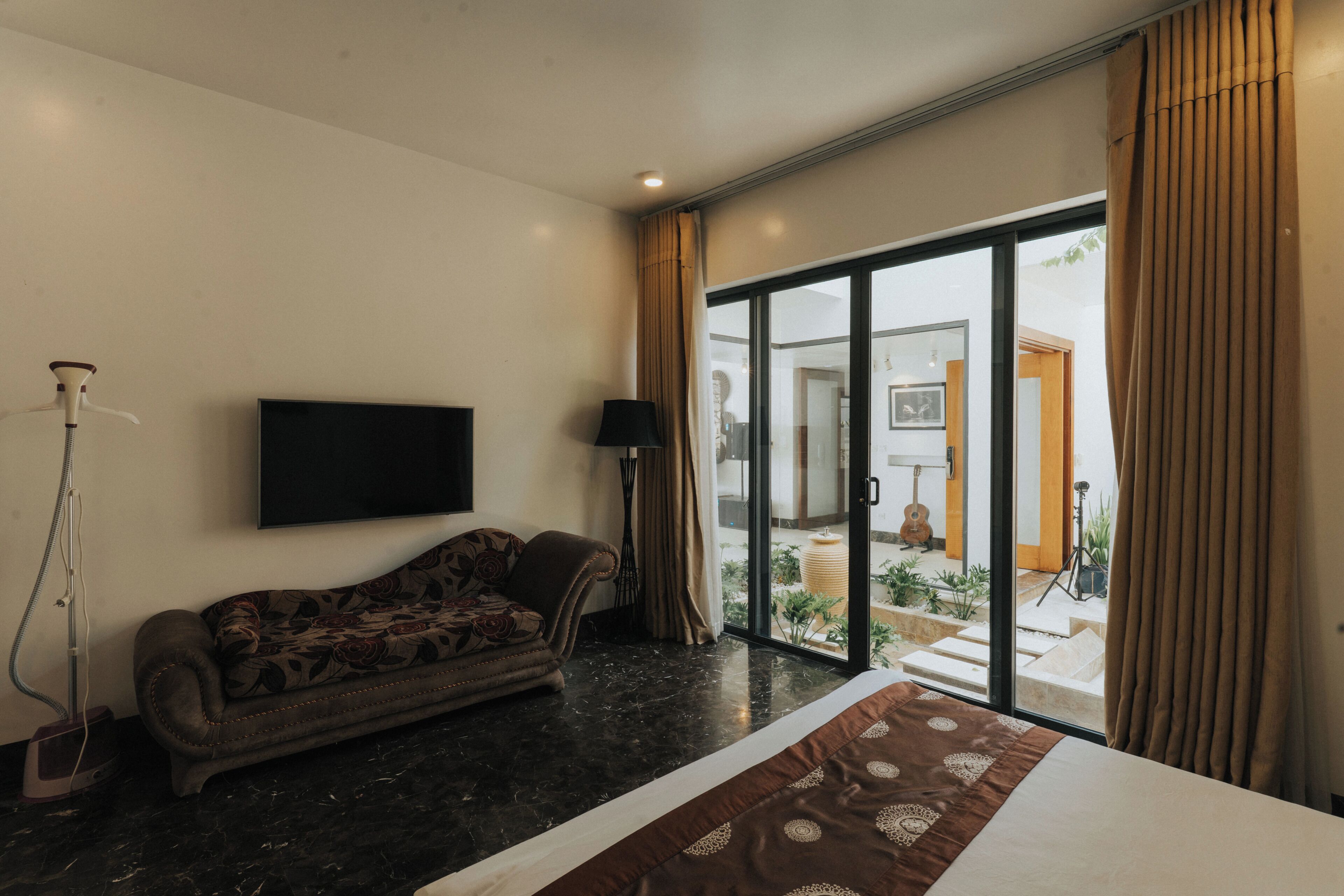 A hotel room with a sofa, a flat-screen TV, a bed, a large sliding glass door leading to an outdoor area, and a floor lamp.