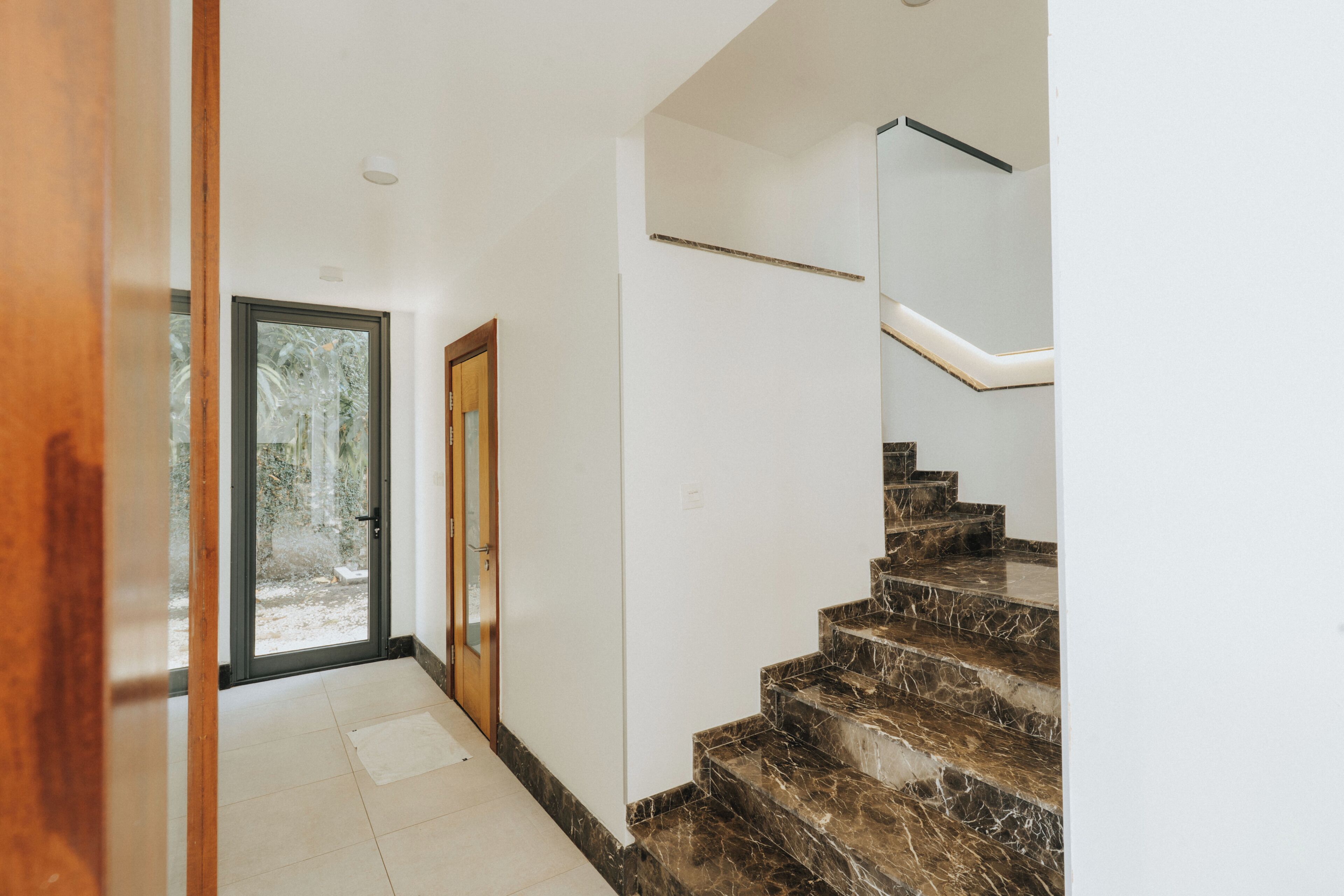 A hallway with a glass door leading outside, a marble staircase, and a white wall.