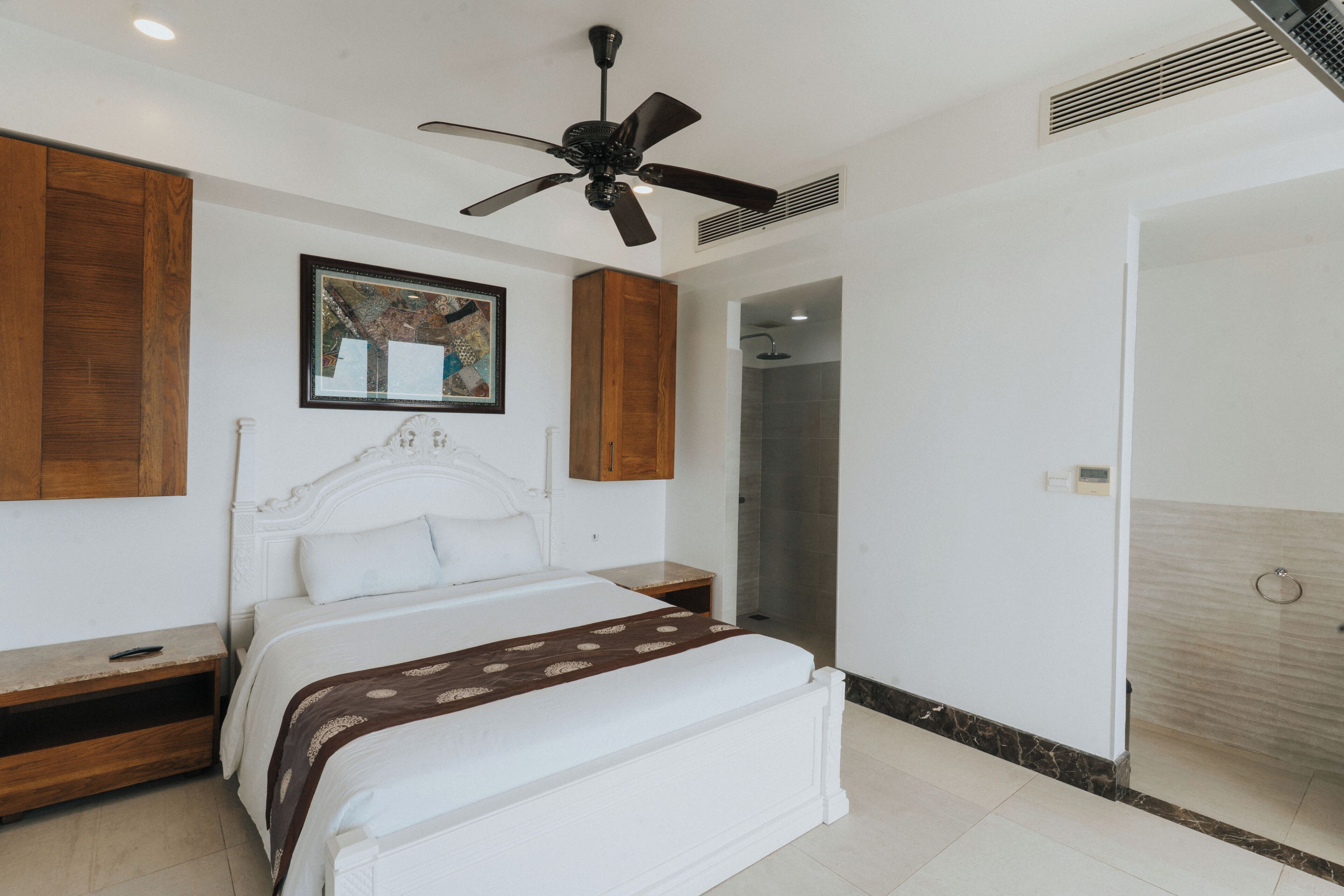 Une chambre à coucher comprenant un lit, une tête de lit en bois, un ventilateur de plafond, un tableau encadré et une salle de bain avec douche.