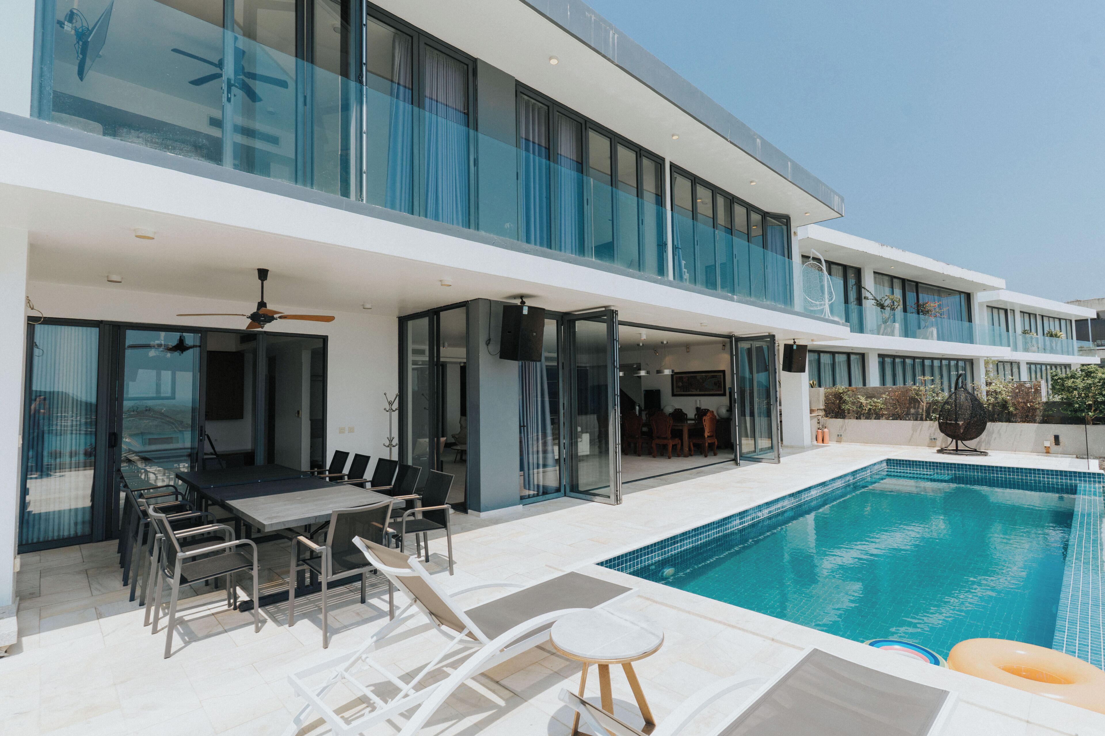 A modern two-story building with a glass facade, a swimming pool, and outdoor seating.