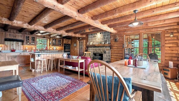 A log cabin interior with a stone fireplace, wooden furniture, and a large window. - White Mountains Cabin w/ Pond & Scenic Deck (Lisbon)