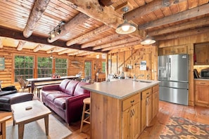A cabin interior with a wooden interior, a kitchen with a stainless steel refrigerator, a dining area with a leather sofa, and a large window. - White Mountains Cabin w/ Pond & Scenic Deck (Lisbon)