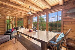 Interior de una cabaña de madera con una gran mesa de comedor, asientos de cuero y ventanas amplias que ofrecen vistas al exterior.