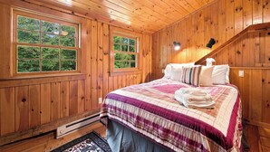 A wooden cabin bedroom with a bed, two windows, a heater, and a staircase. - White Mountains Cabin w/ Pond & Scenic Deck (Lisbon)