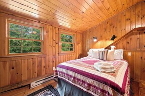 Un dormitorio de cabaña de madera con una cama, dos ventanas, un calentador y una escalera.