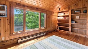 A wooden cabin interior with large windows, a built-in bookshelf, and a heating unit. - White Mountains Cabin w/ Pond & Scenic Deck (Lisbon)