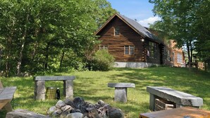 A rustic log cabin with a stone fire pit and benches in a lush green setting. - White Mountains Cabin w/ Pond & Scenic Deck (Lisbon)