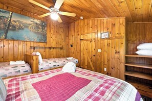 Una habitación tipo cabaña con dos camas, un ventilador de techo, un cuadro de un paisaje y una estantería de madera con almohadas.