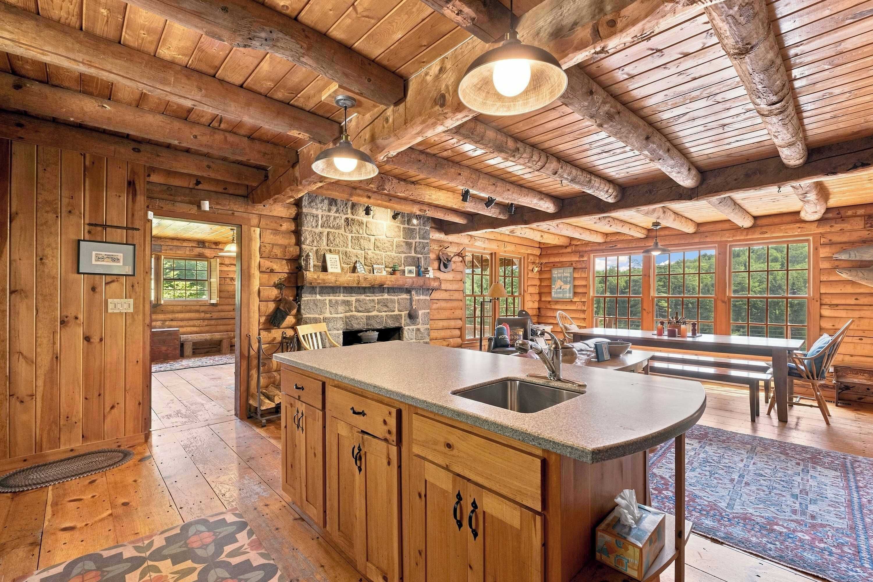 Interior de una cabaña de madera con zona de cocina, chimenea de piedra y amplios ventanales.