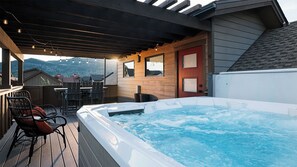 A hot tub on a deck with chairs, a wooden pergola, and a view of mountains.