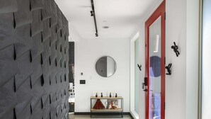 Un pasillo con pared texturizada, un espejo redondo, un banco con almohadones decorativos y una puerta con marco rojo.