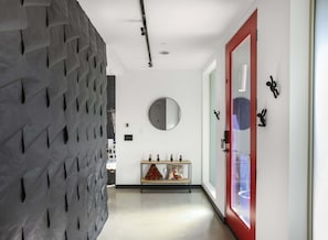 A hallway with a textured wall, a round mirror, a bench with decorative pillows, and a red-framed door. - Welcome to Casa Black - 3 Bedroom 3 Bathroom in the Palm Springs Area (Palm Springs)