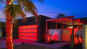 Un edificio moderno con una puerta de garaje iluminada en rojo, un patio cubierto y una palmera.