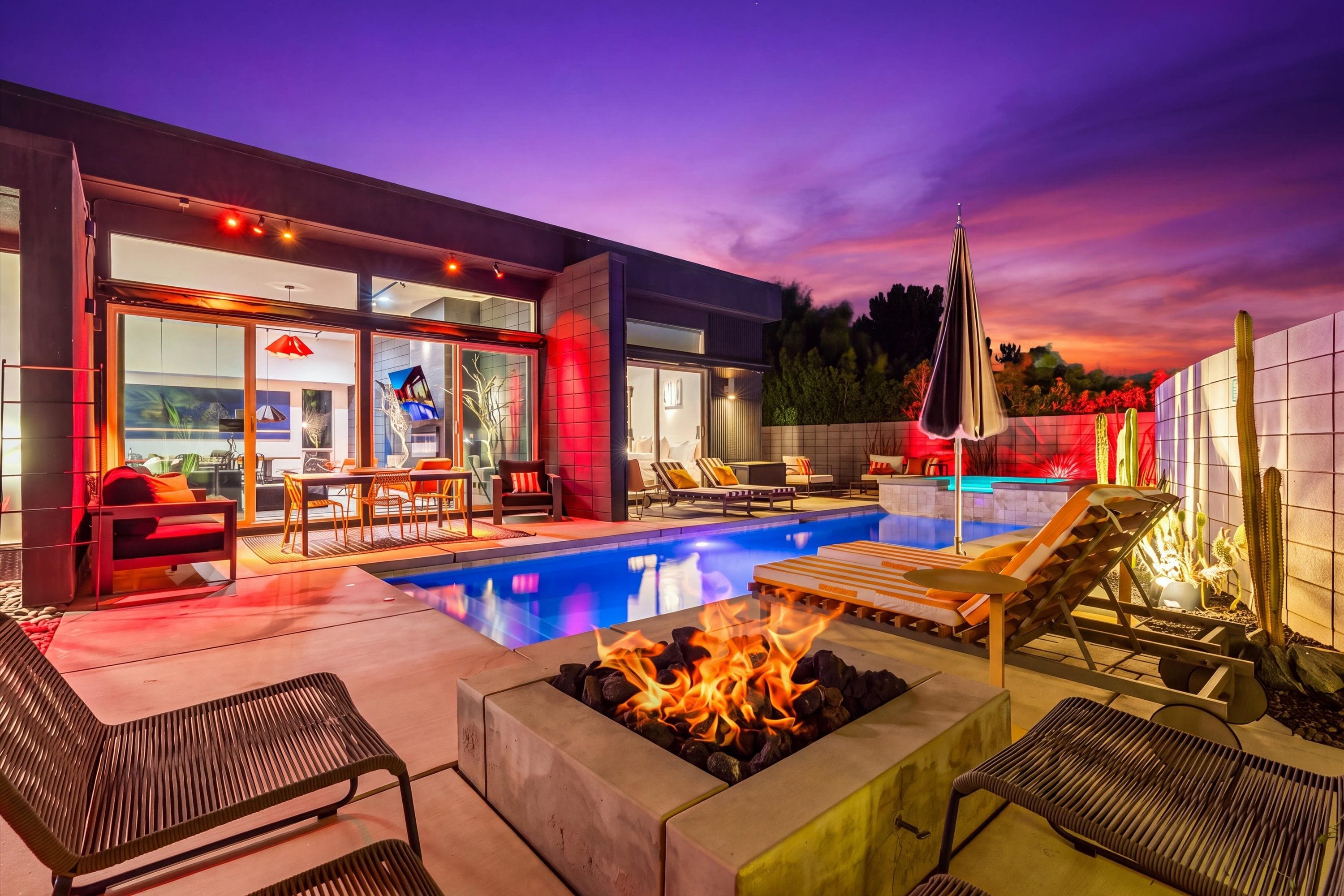 Een modern zwembad met ligbedden, een vuurkorf en een patio met glazen wanden.