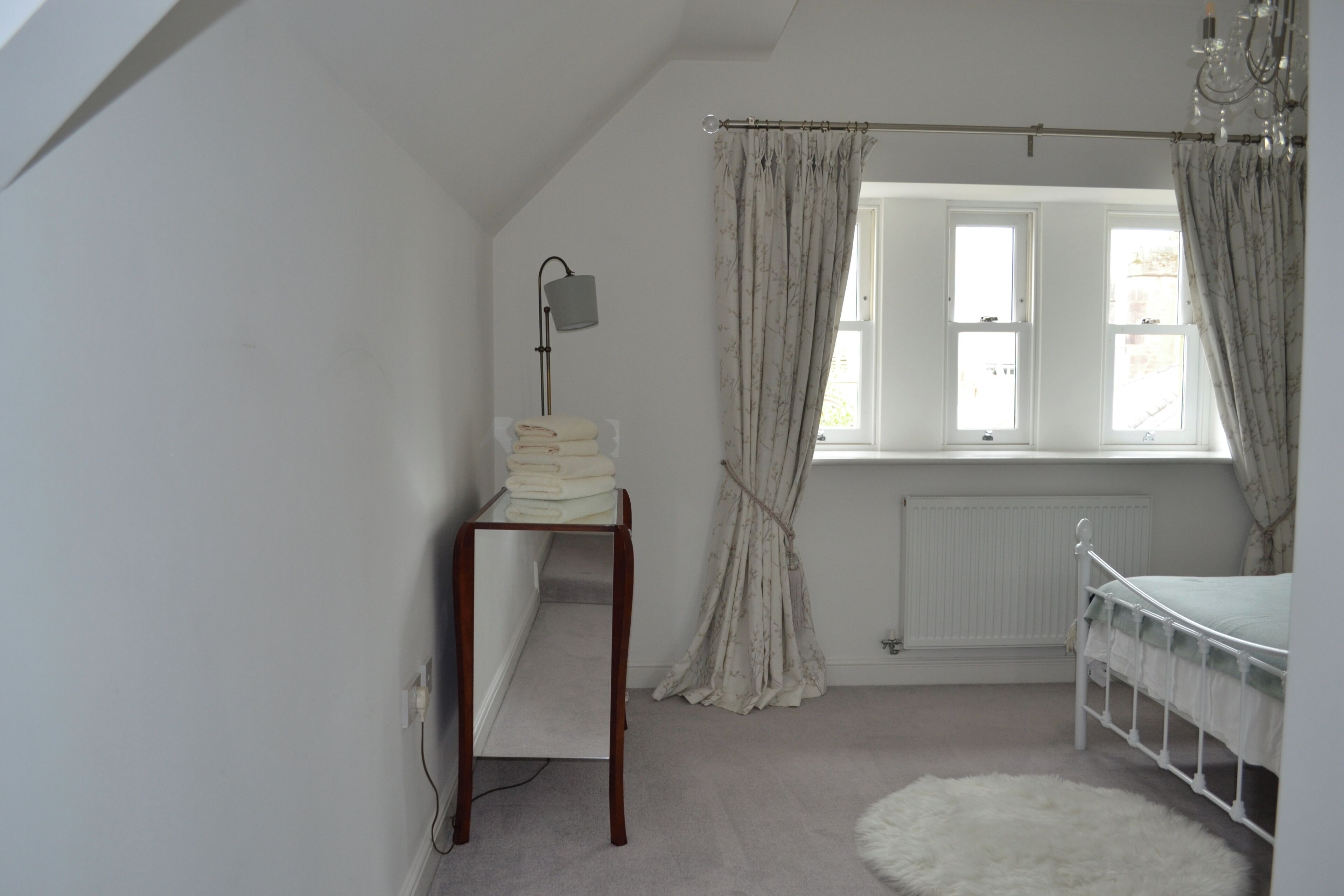 A bedroom with a window, a radiator, a bedside table with a lamp, and a white fluffy rug.